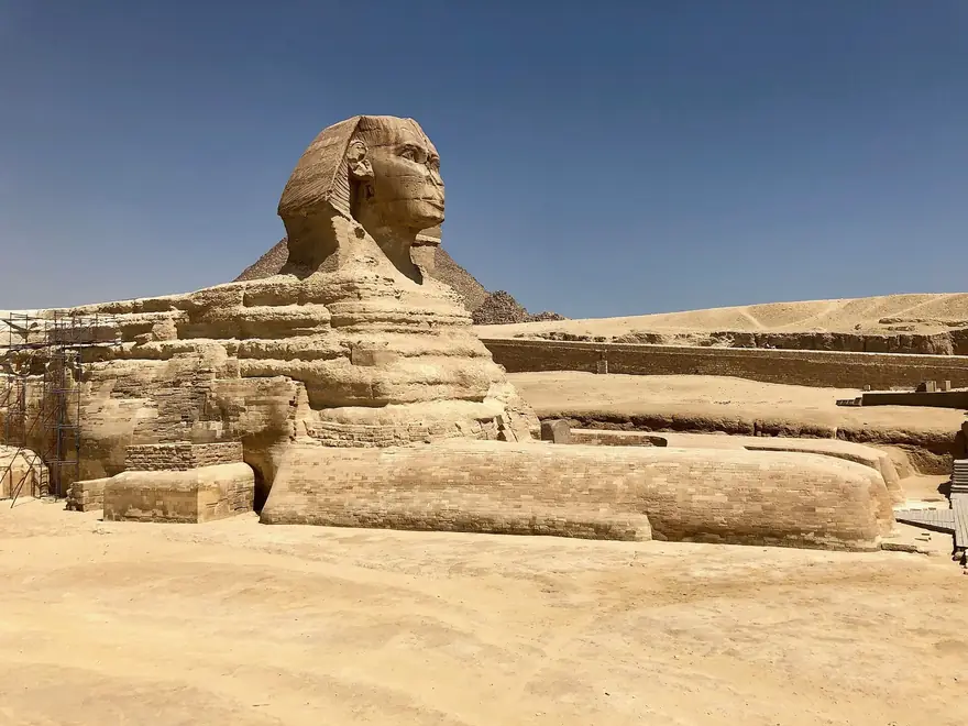 Die Große Sphinx von Gizeh unter blauem Himmel, im Hintergrund eine Pyramide